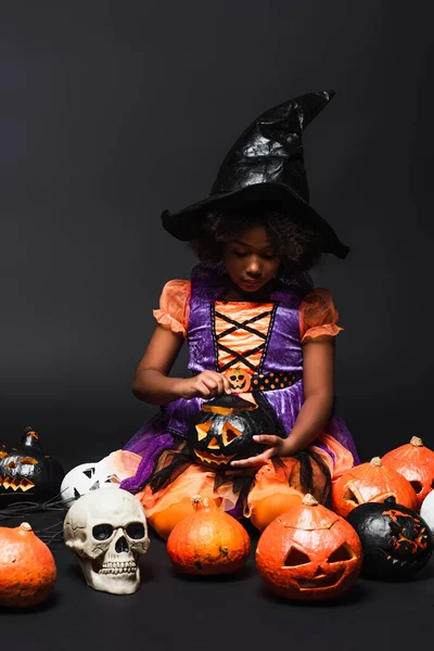 African american girl in witch costume and pointed hat looking at carved pumpkin on black — Stock Photo