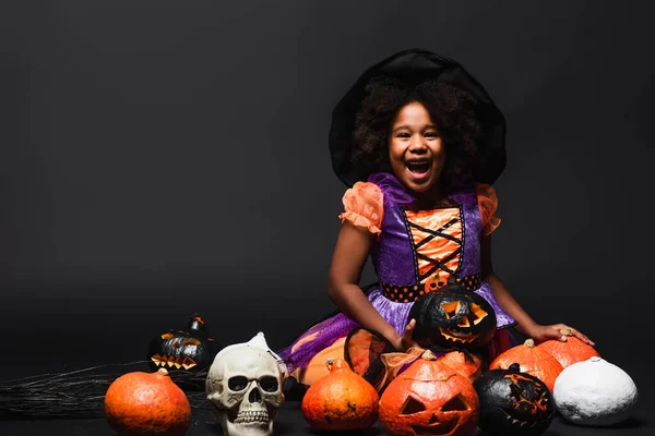 Excitada chica afroamericana en traje de bruja y sombrero puntiagudo cerca de calabazas talladas en negro - foto de stock