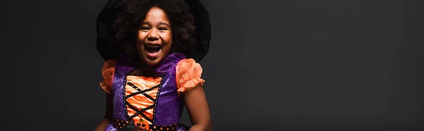 Cheerful african american girl in witch costume and pointed hat isolated on black, banner — Stock Photo