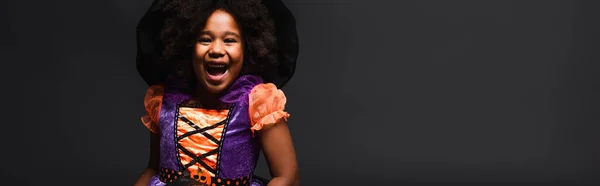 Alegre afroamericana chica en traje de bruja y sombrero puntiagudo aislado en negro, bandera - foto de stock