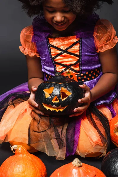 Cheerful african american girl in costume holding carved pumpkin isolated on black — Stock Photo