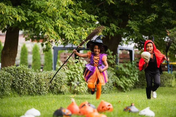 Crianças africanas americanas em trajes de Halloween segurando vassouras e correndo no quintal — Fotografia de Stock