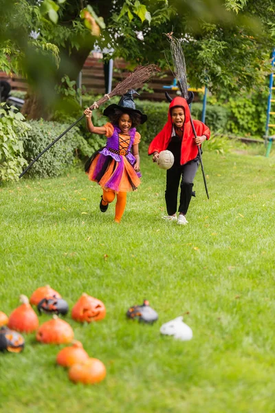 Возбужденные африканские американские дети в костюмах на Хэллоуин держат метлы и бегают по заднему двору — стоковое фото