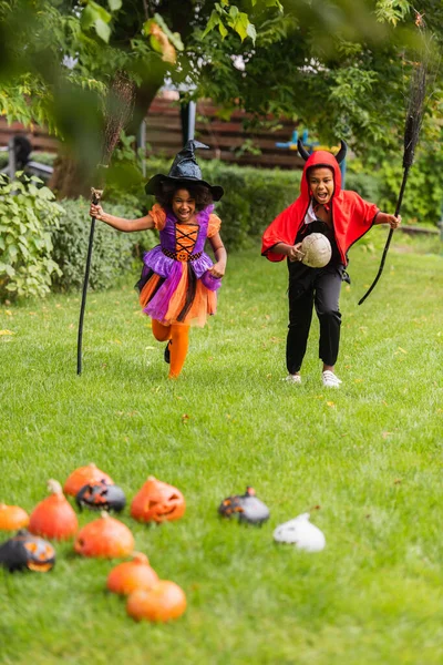 Возбужденные африканские американские дети в костюмах на Хэллоуин держат метлы и бегают по газону — стоковое фото