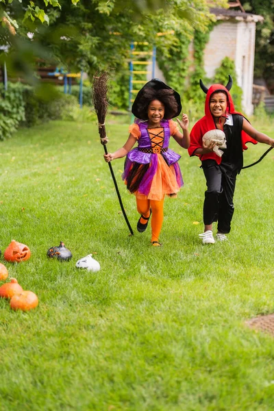 Felici bambini afro-americani in costumi di Halloween che tengono scope e corrono sul prato — Foto stock