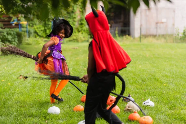 Frères et sœurs afro-américains en costumes d'Halloween tenant des balais et jouant sur la pelouse — Photo de stock