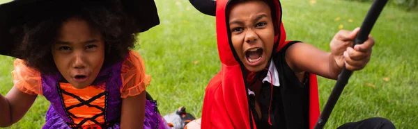 African american siblings in halloween costumes holding brooms and playing on backyard, banner — Stock Photo