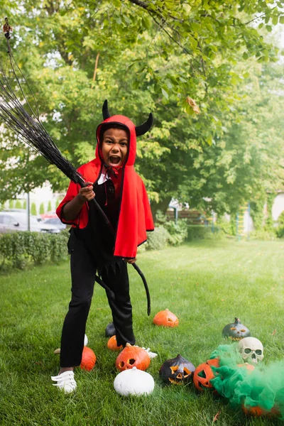 Scary african american boy in devil halloween costume holding broom near pumpkins on lawn — Stock Photo