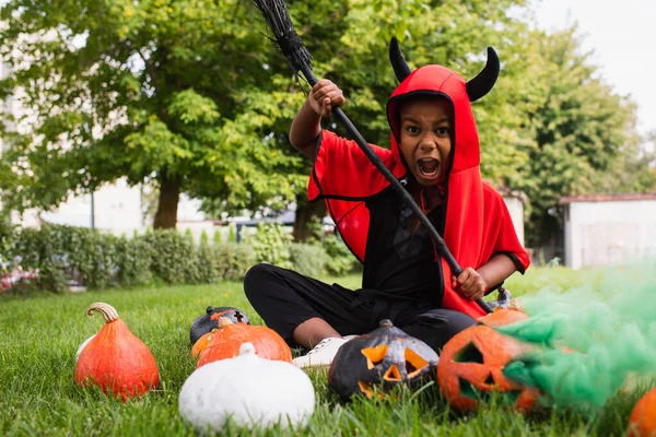 Gruseliges afrikanisch-amerikanisches Kind im Teufel-Halloween-Kostüm schreit, während es Besen in der Nähe von Kürbissen hält und auf dem Rasen sitzt — Stockfoto