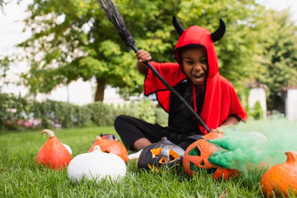 Borrosa afroamericano chico en diablo halloween traje gritando mientras sostiene escoba cerca de calabazas y sentado en el césped - foto de stock