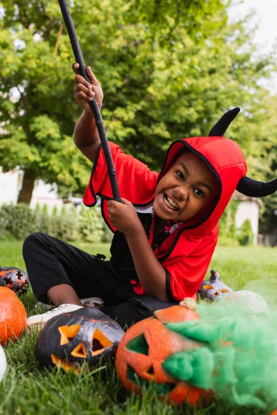 Offuscata africano americano ragazzo in diavolo halloween costume ghigno mentre tenendo scopa vicino zucche e seduto sul prato — Foto stock