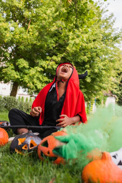 Spaventoso africano americano bambino in diavolo halloween costume ridere forte mentre tenendo scopa vicino zucche e seduto sul prato — Foto stock