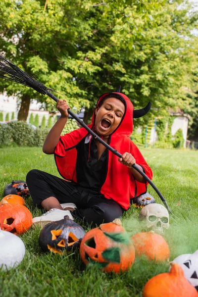 Wütender afrikanisch-amerikanischer Junge im Teufel-Halloween-Kostüm schreit, während er Besen in der Nähe von Kürbissen hält und auf dem Rasen sitzt — Stockfoto