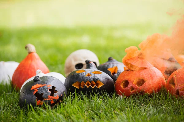 Carved and scary pumpkins with orange smoke near blurred skull on green lawn — Stock Photo