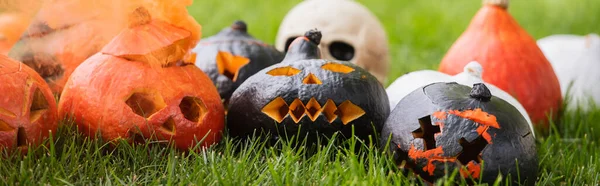 Carved and scary pumpkins with orange smoke near blurred skull on green lawn, banner — Stock Photo