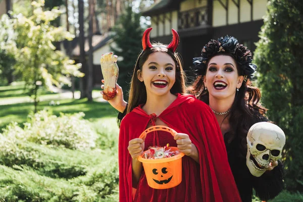 Animado mãe e filha no diabo e vampiro halloween trajes, com crânio, mão de brinquedo e balde de doces — Fotografia de Stock