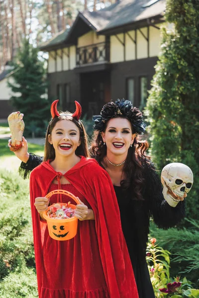 Cheerful girl and mother in halloween costumes, with bucket of sweets, skull and toy hand in backyard — Stock Photo