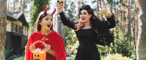 Frau im Vampir-Halloween-Kostüm erschreckt Tochter mit Spielzeughand und Totenkopf, Banner — Stockfoto