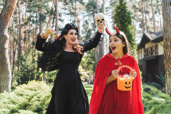 Chica en diablo halloween traje celebración cubo con caramelos y gritando cerca de mamá con cráneo y juguete mano - foto de stock