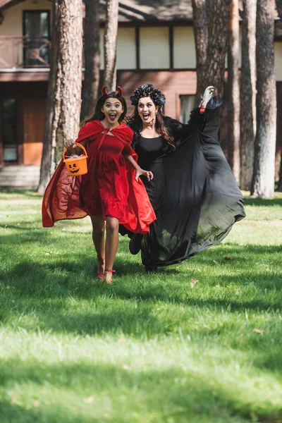 Excited woman and girl in halloween costumes screaming while running in backyard — Stock Photo