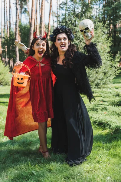 Mutter und Tochter in Halloween-Kostümen halten Eimer mit Süßigkeiten, Totenkopf und Spielzeughand, während sie im Wald schreien — Stockfoto