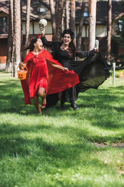 Emocionado mamá y su hija en trajes de halloween corriendo con cráneo, mano de juguete y cubo de caramelos - foto de stock