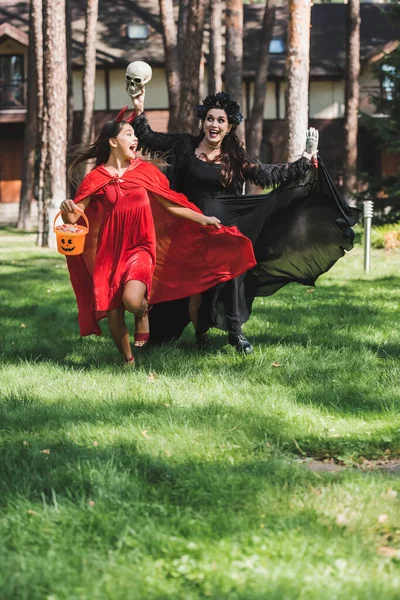 Eccitati mamma e figlia in costumi di Halloween in esecuzione con teschio, mano giocattolo e secchio di caramelle — Foto stock