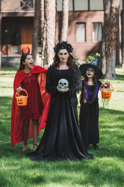 Mujer en traje de halloween vampiro sosteniendo cráneo cerca de hijas espeluznantes con cubos de dulces - foto de stock