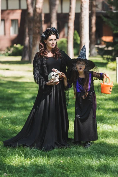 Mujer en traje de halloween vampiro cerca de hija en sombrero de bruja mueca mientras sostiene cubo con caramelos - foto de stock