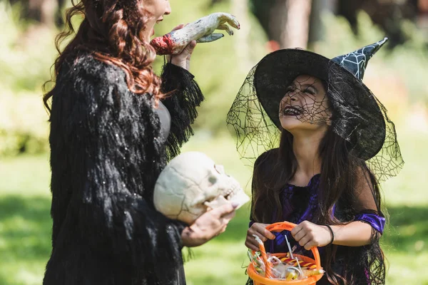 Mujer borrosa en traje de halloween negro asustando hija alegre con mano de juguete - foto de stock