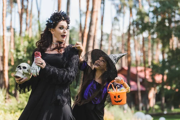 Niño en sombrero de bruja apuntando a cubo de Halloween con caramelos cerca de mamá emocionada en traje de vampiro - foto de stock