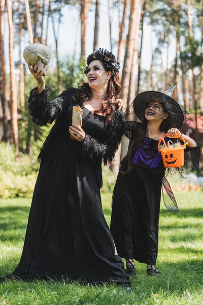 Счастливые мама и дочь в черных костюмах Хэллоуина, с черепом, игрушечной рукой и ведром с конфетками — стоковое фото