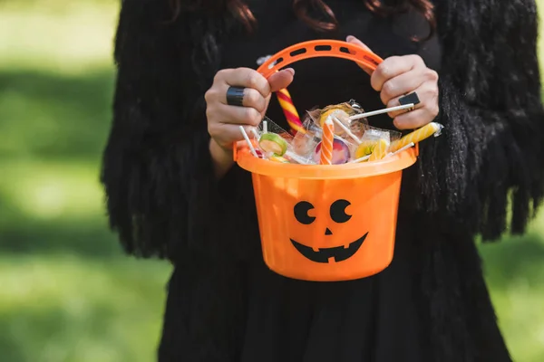 Cropped view of woman in black dress holding halloween bucket with sweets — Stock Photo
