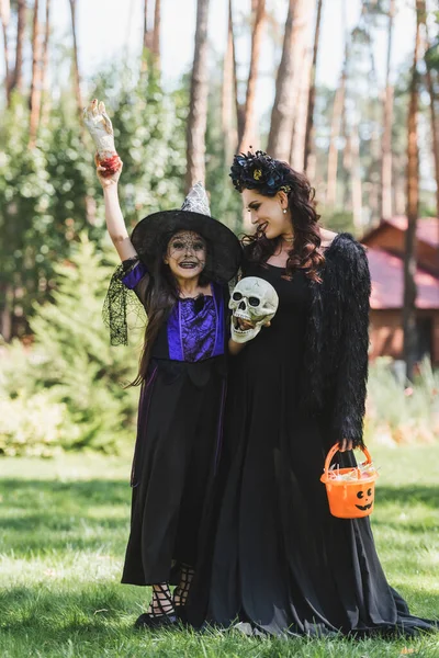 Excited girl in witch halloween costume holding toy hand and skull near mom — Stock Photo