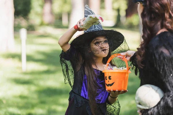 Girl in witch hat holding bucket with candies and scaring mom with toy hand — Stock Photo
