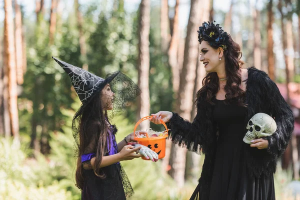 Happy mother and daughter in halloween costumes, with bucket of sweets and skull in forest — Stock Photo