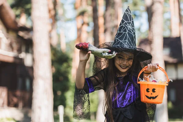 Cheerful girl in witch halloween costume holding toy hand and bucket with sweets outdoors — Stock Photo