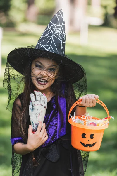 Chica en sombrero de bruja haciendo mueca de miedo mientras sostiene la mano de juguete y cubo con caramelos - foto de stock