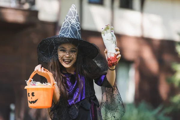 Excitada chica vistiendo disfraz de bruja halloween mientras sostiene juguete de la mano y cubo con dulces - foto de stock