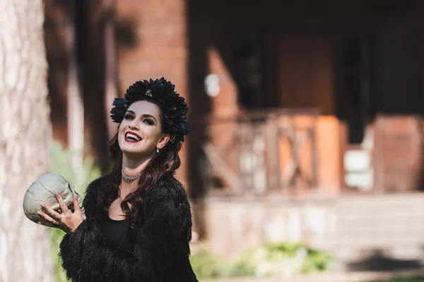 Cheerful woman in vampire halloween costume holding spooky skull in backyard — Stock Photo