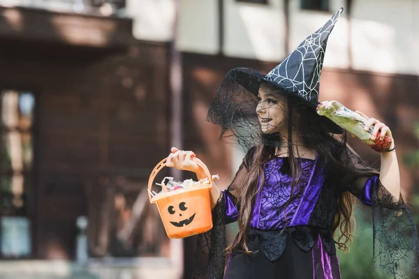 Happy child in witch halloween costume, with toy hand and bucket with candies looking away outdoors — Stock Photo