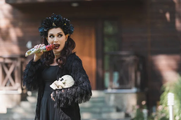 Mulher em vampiro halloween traje segurando crânio e mordendo brinquedo mão ao ar livre — Fotografia de Stock
