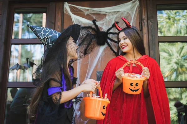 Happy girls in halloween costumes holding buckets of candies on house porch with decoration — Stock Photo