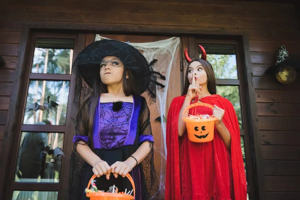 Chica en traje del diablo mostrando signo de silencio cerca ofendida hermana en sombrero de bruja - foto de stock