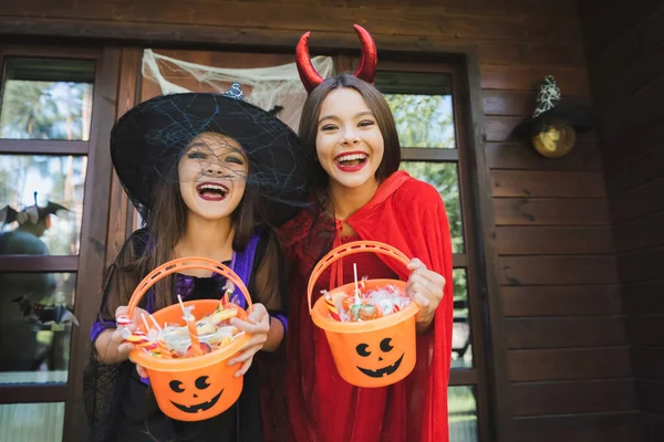 Excited sisters in witch and devil halloween costumes holding buckets with sweets — Stock Photo