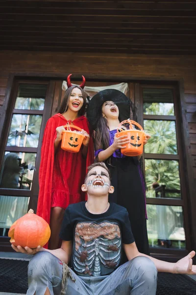 Crianças animadas em fantasias assustadoras de Halloween segurando baldes com doces e gritando no alpendre — Fotografia de Stock