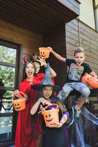 Kinder in gruseligen Halloween-Kostümen zeigen gruselige Gesten, während sie Eimer mit Süßigkeiten auf der Veranda halten — Stockfoto