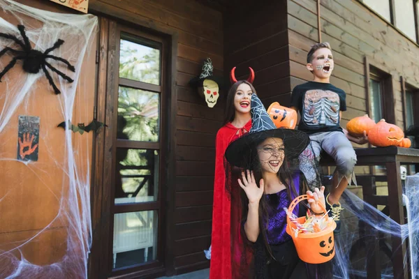 Chica en sombrero de bruja agitando la mano cerca de niños emocionados en trajes espeluznantes de halloween en el porche con decoración - foto de stock