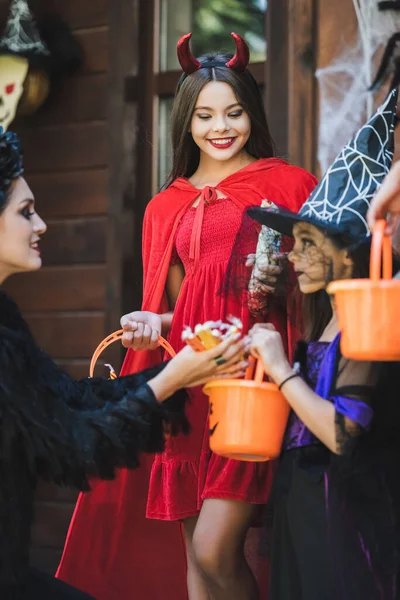 Mom giving candies to happy daughters in devil and witch halloween costumes — Stock Photo