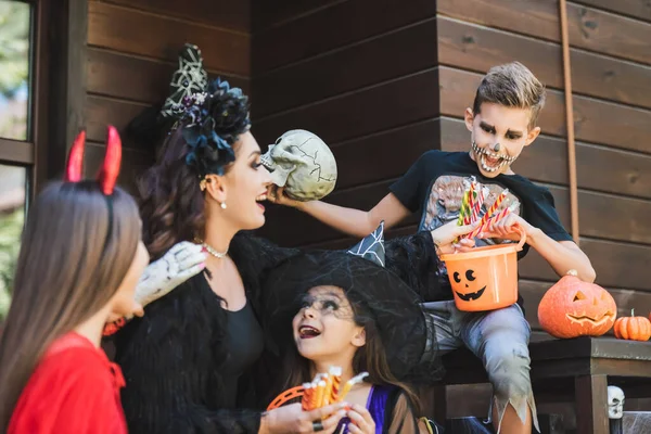 Niño sosteniendo cubo con caramelos cerca asombrado mamá y hermanas en trajes de halloween - foto de stock